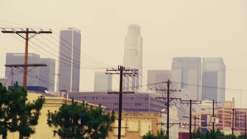 A slow-motion driving shot reveals the hazy skyline of downtown Los Angeles behind trees and power lines