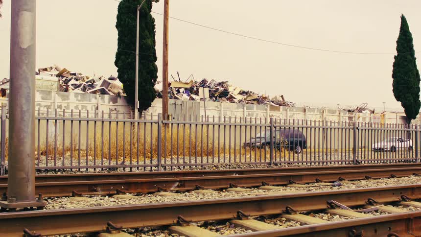 A metro train travels on railroad tracks past a junkyard in an industrial part of the city