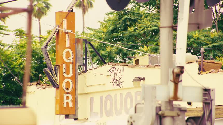 A vintage-style shot shows an old, weathered liquor store sign next to a railroad crossing as cars drive by in an urban setting