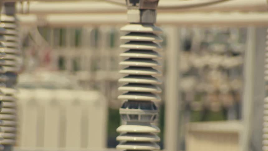 A rack focus shot of high-voltage insulators and a transformer at an electrical substation on a hot day