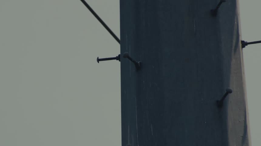 An extreme close-up slow tilt down a high-voltage power line tower, showing insulators and metal details against a flat grey sky