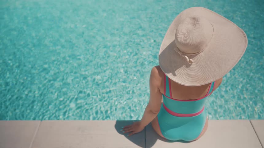 A woman in a stylish swimsuit and wide-brimmed hat relaxes by the edge of a sparkling blue swimming pool on a sunny day