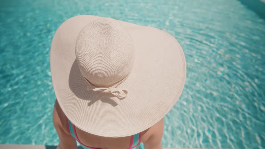 A woman wearing a large, elegant sun hat relaxes at the edge of a shimmering turquoise swimming pool