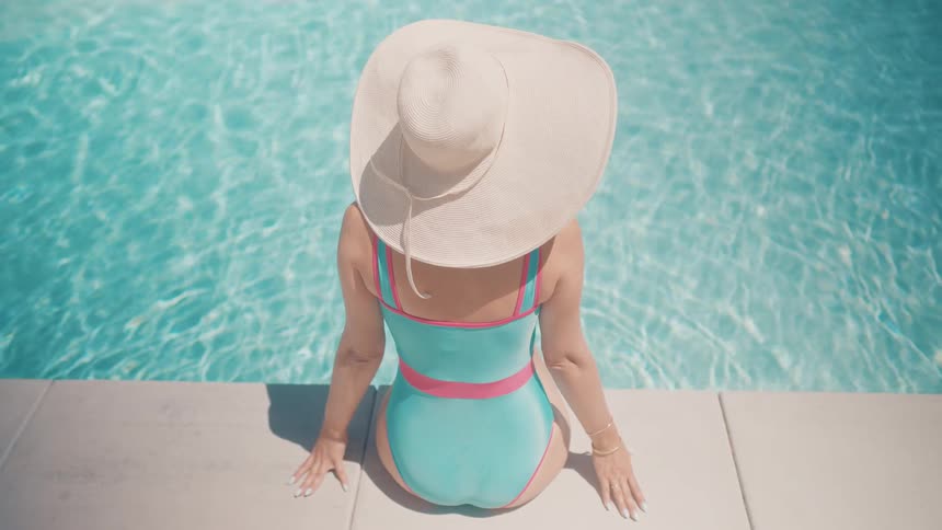 High-angle view from behind of a stylish woman in a swimsuit and hat relaxing by the pool on a sunny day