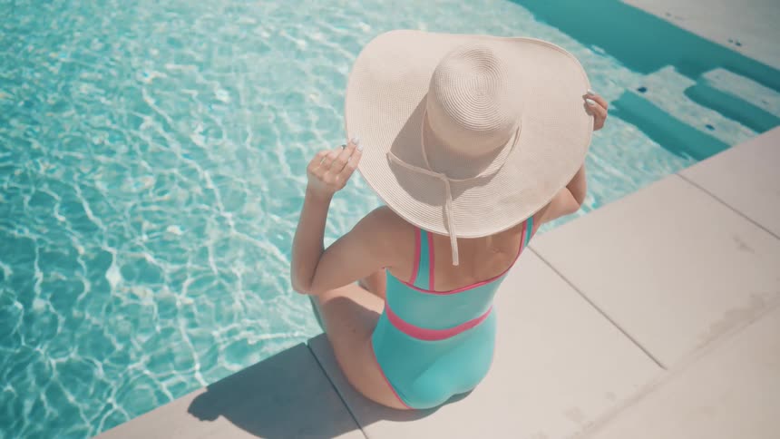 A woman in a stylish swimsuit and wide-brimmed hat relaxes by the edge of a crystal clear swimming pool on a sunny day