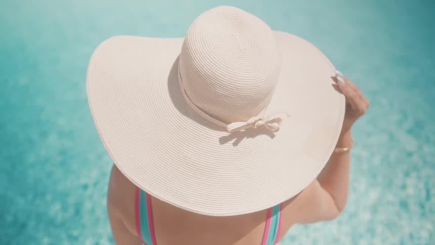 A high-angle view shows a woman in a large straw hat relaxing by the turquoise water of a swimming pool on a sunny day