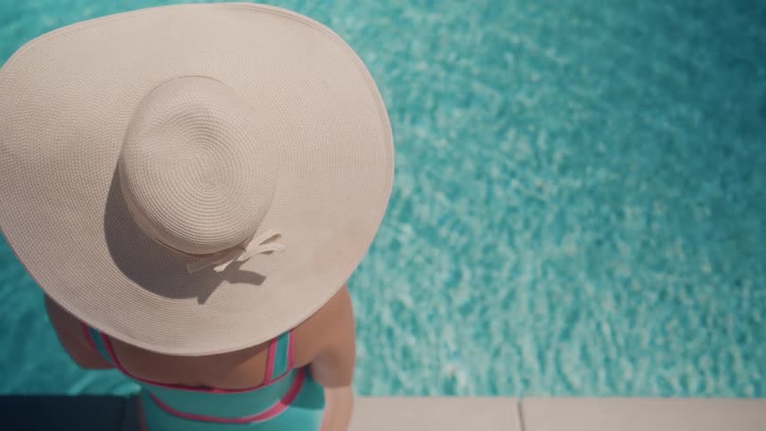 A woman in a large sun hat and colorful swimsuit relaxes by the edge of a sparkling blue swimming pool on a sunny day