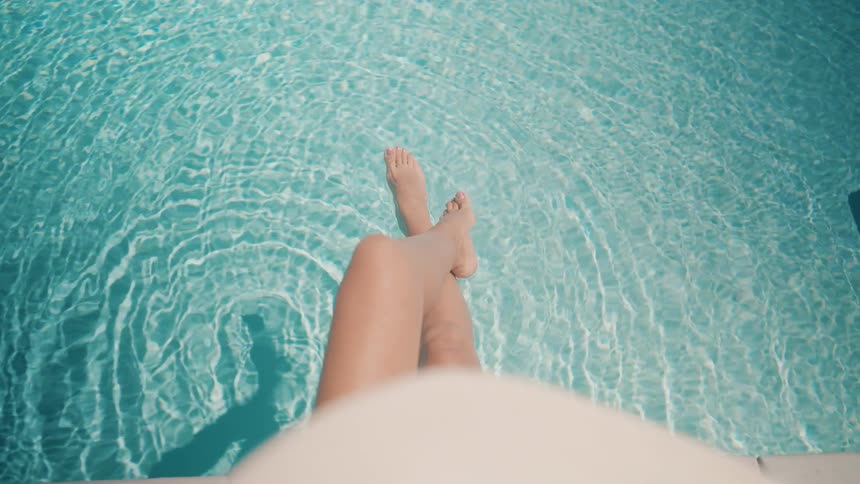 A first-person view of a woman's legs relaxing in the cool, rippling water of a sunlit swimming pool