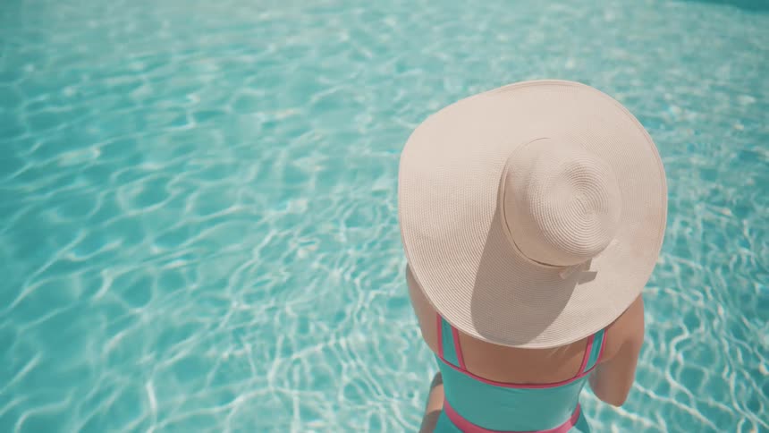 A woman in a wide-brimmed straw hat sits relaxing in the shimmering turquoise water of a swimming pool on a sunny day