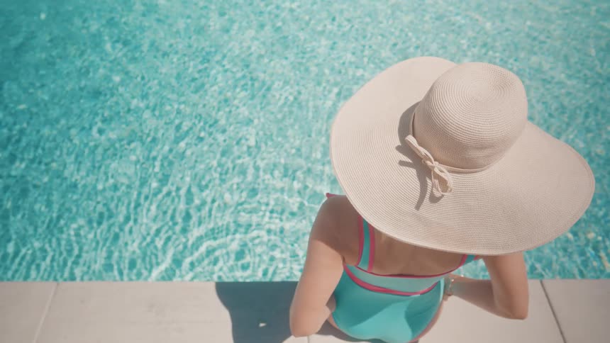 Overhead view of a woman in a floppy hat enjoying a relaxing sunny day by the clear blue water of a swimming pool