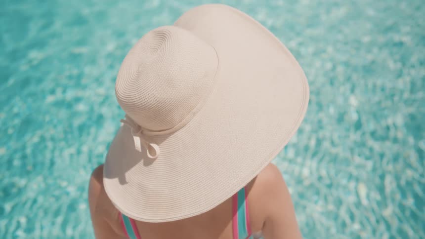 High angle view of a woman in a large straw hat relaxing in a bright turquoise swimming pool on a sunny summer day