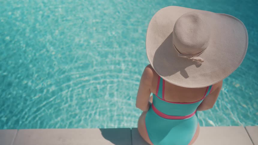 A woman in a stylish swimsuit and wide-brimmed hat relaxes by the edge of a sparkling swimming pool on a sunny day