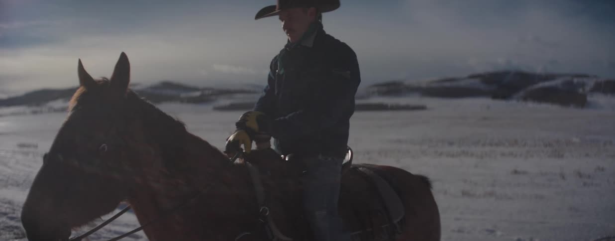 A lone cowboy sits stoically on his horse in a vast, snow-covered field as the winter sun creates a bright lens flare