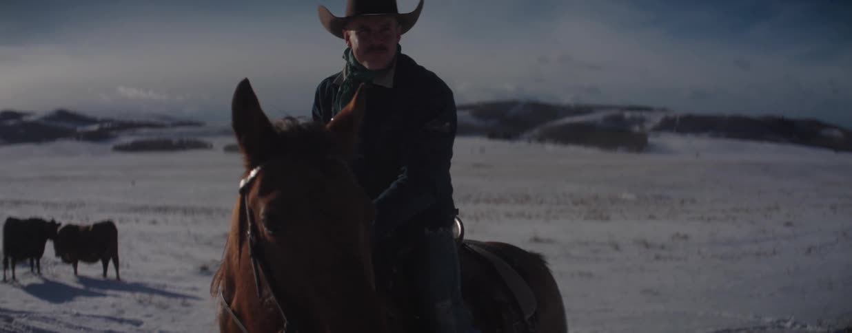 A cowboy on horseback sits stoically in a vast, snowy field as the camera slowly moves past him, with a herd of cattle visible in the background