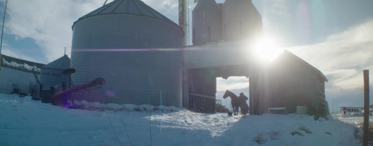 A cowboy prepares his horse in the opening of a barn on a snowy farm, silhouetted by the bright winter sun next to large silos