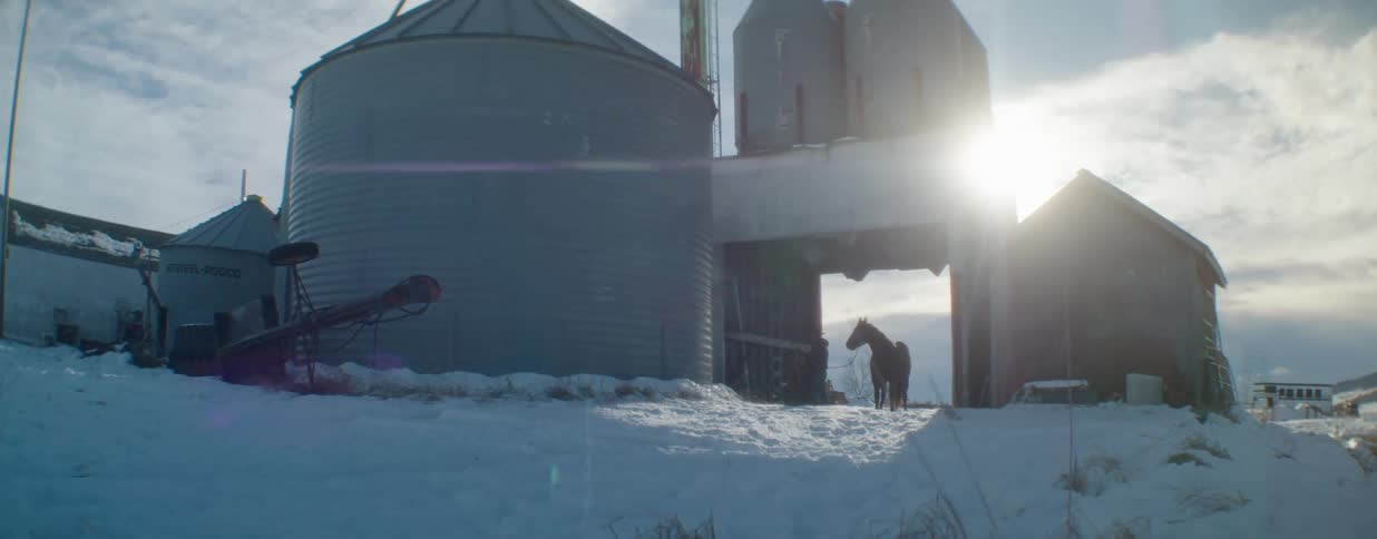 A silhouetted rancher leads a horse from a snowy barn at sunrise next to large grain silos