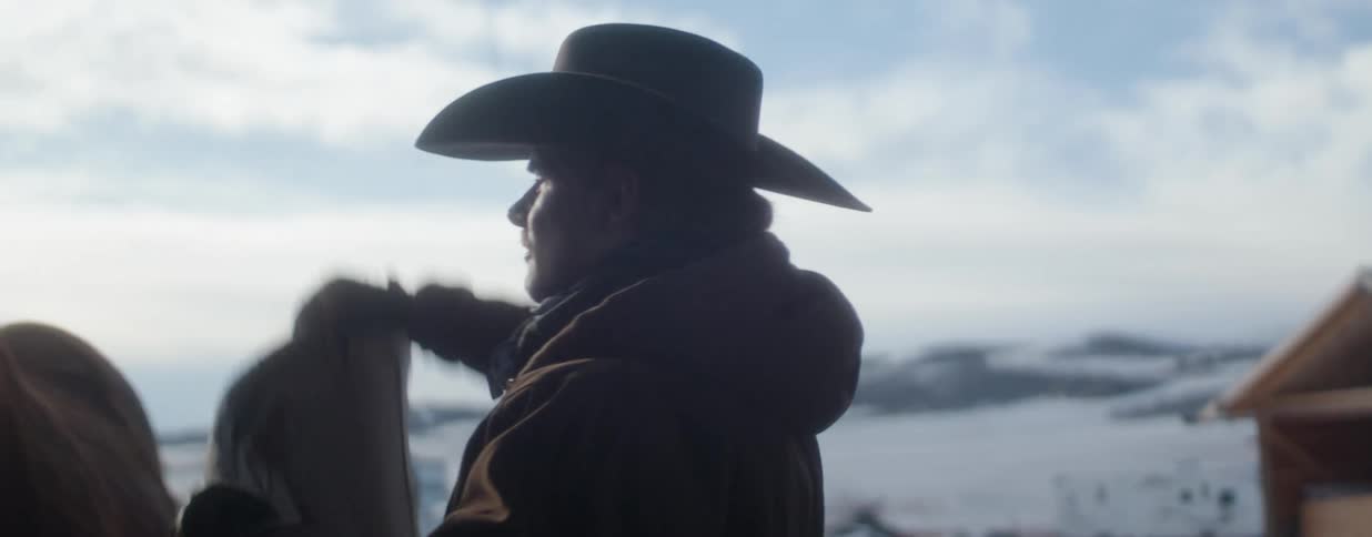 A rancher in a cowboy hat saddles his horse in a barn, silhouetted against a snowy winter landscape