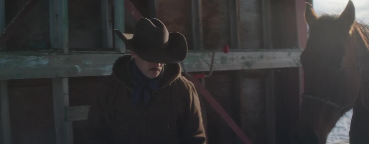A cowboy in a winter coat leads his brown horse out of the snow and into a rustic wooden barn