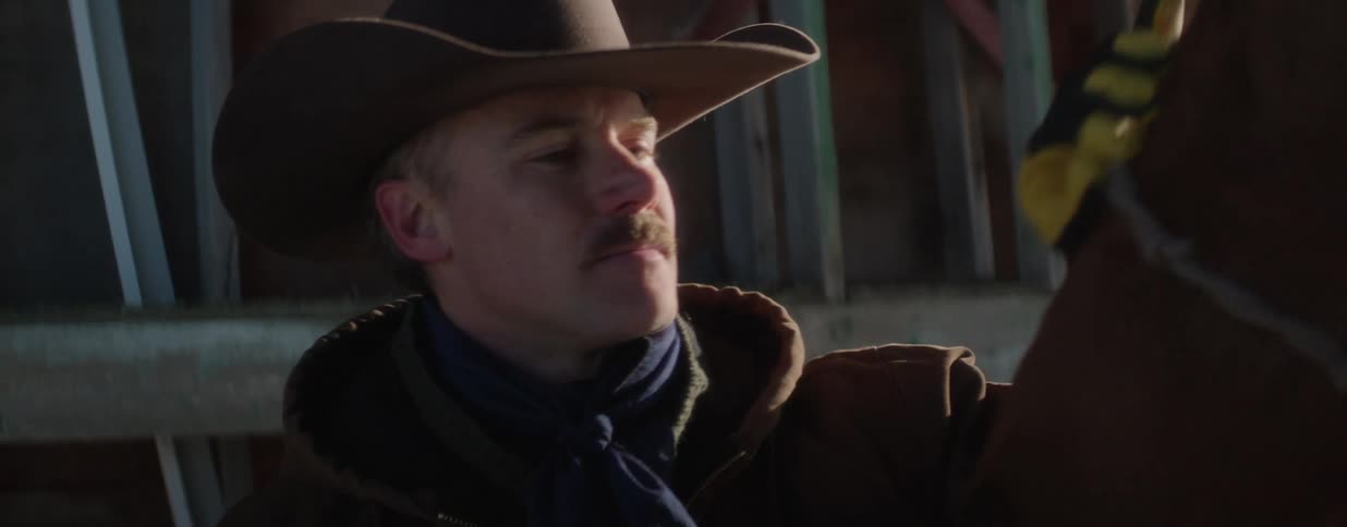 A thoughtful cowboy stands beside his horse in a rustic barn, bathed in the warm glow of sunlight