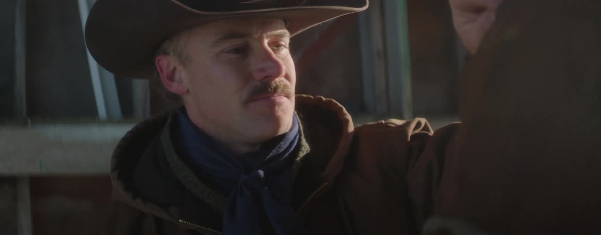 A kind cowboy with a mustache gently pets his horse in a sunlit barn