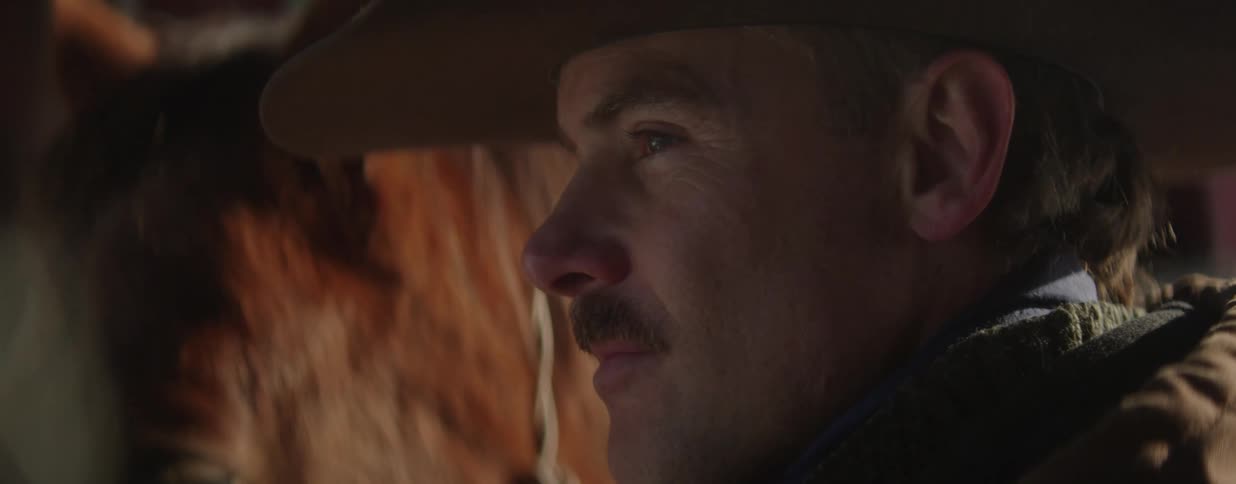A thoughtful cowboy wearing a hat rests his eyes in a moody, dimly lit profile close-up