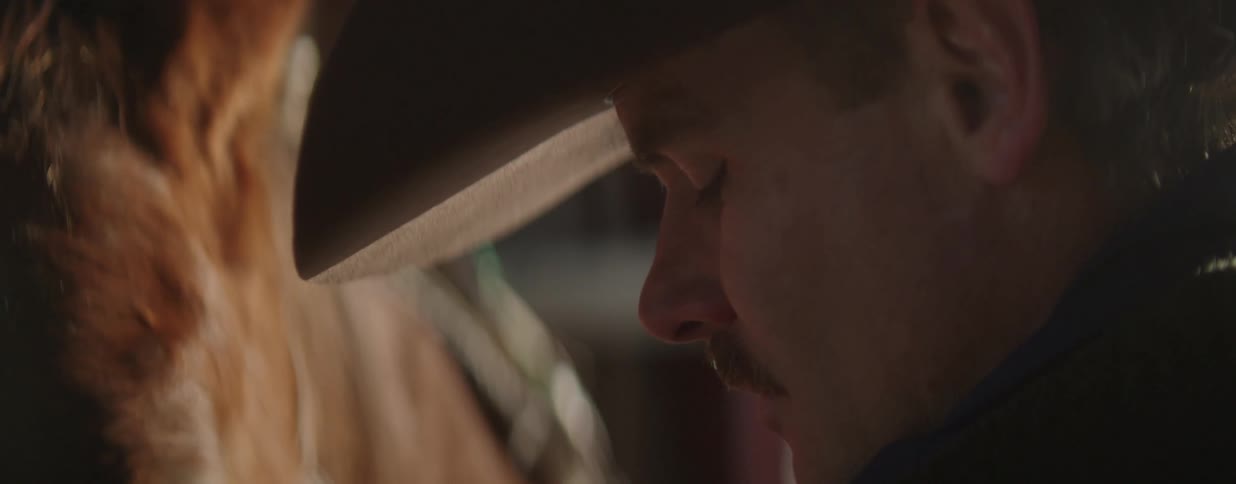 A cowboy wearing a hat has a quiet, contemplative moment with his eyes closed next to his horse in a stable