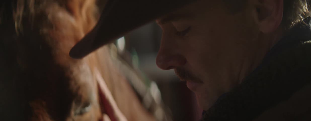 An intimate close-up of a cowboy with his eyes closed, gently stroking his horse in a dimly lit barn