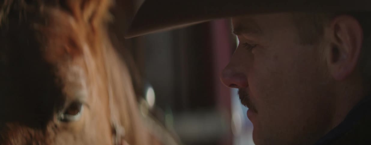 An intimate close-up shows a cowboy gently stroking his horse's face inside a barn