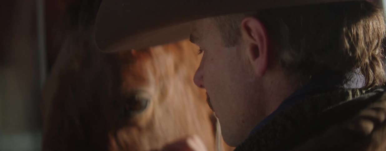 A cowboy in a dimly lit stable gently caresses the side of his brown horse's face