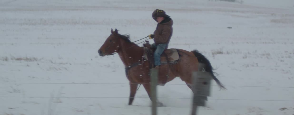 A cowboy on horseback gallops across a vast, snow-covered landscape in dramatic slow motion