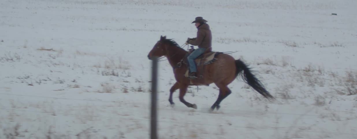 A man in a cowboy hat gallops on his horse across a vast, snow-covered plain in slow motion