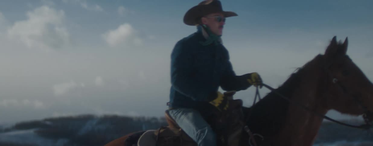 A rugged cowboy in a hat and sunglasses rides his horse in slow motion across a vast, snowy landscape