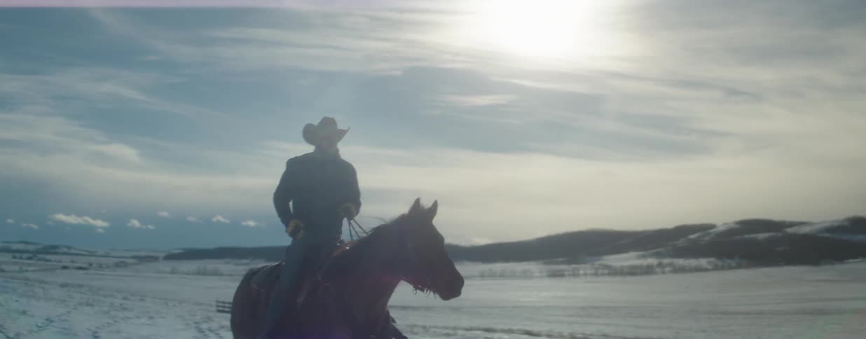 A cowboy rides his horse in slow motion across a vast and beautiful snow-covered landscape under a bright, cold sun