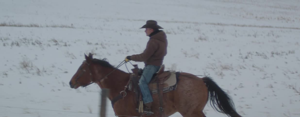 Slow motion side profile of a cowboy riding his galloping horse through a snowy winter landscape