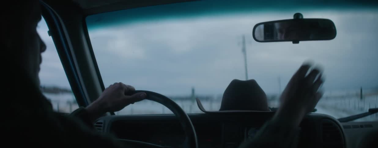 A man adjusts his rearview mirror while driving his truck down a desolate country road on a cold, overcast day