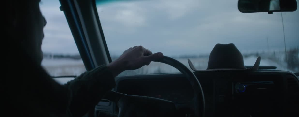 Over-the-shoulder shot from inside a truck of a person driving through a snowy, rural landscape with a cowboy hat on the dashboard