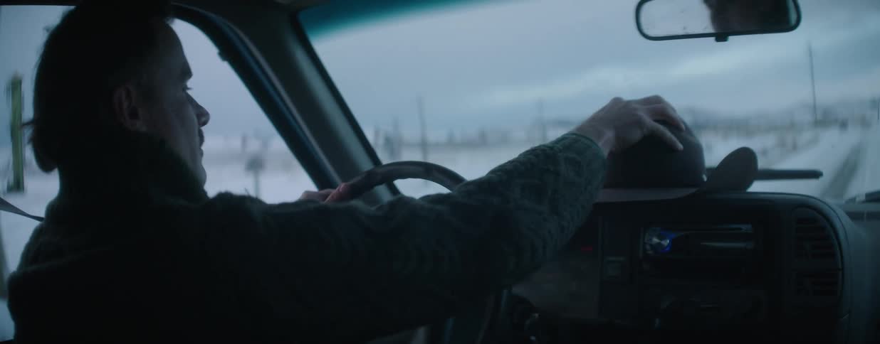 A mustached man in a green sweater drives his pickup truck through a bleak, snowy countryside in slow motion