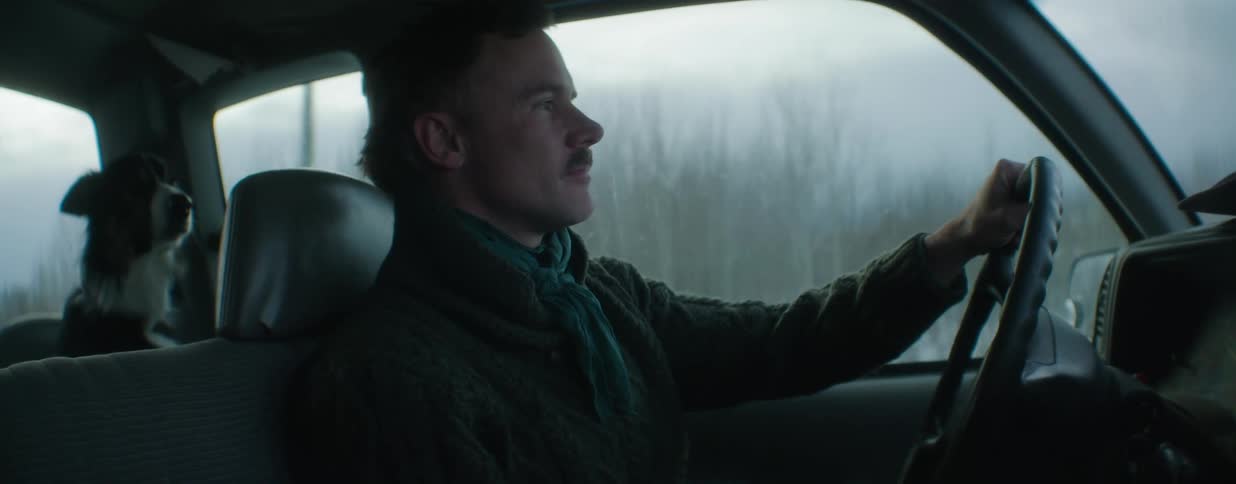 A man with a mustache wearing a cozy sweater drives his car on a cool, overcast day