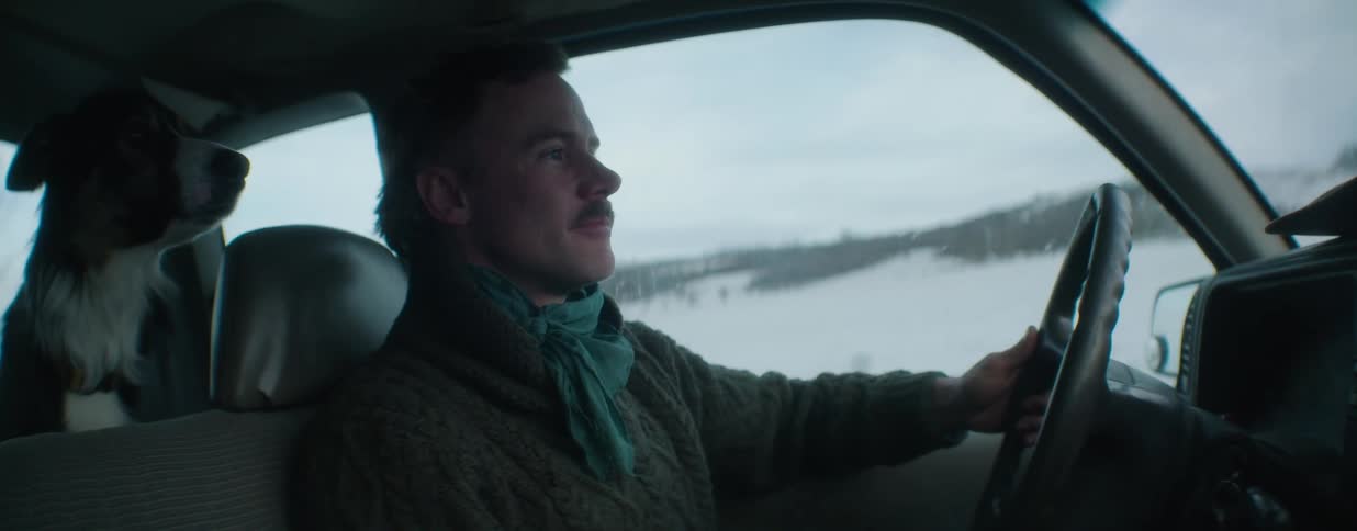 A content man with a mustache drives his truck through a snowy, desolate landscape with his dog in the passenger seat