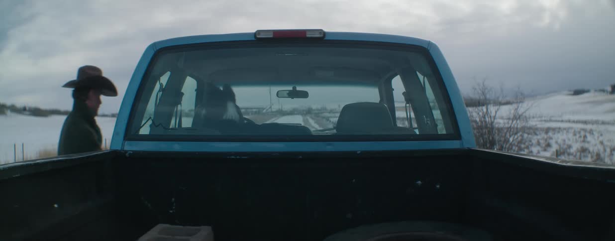 A cowboy gets into his pickup truck with his dog waiting in the passenger seat on a snowy rural road