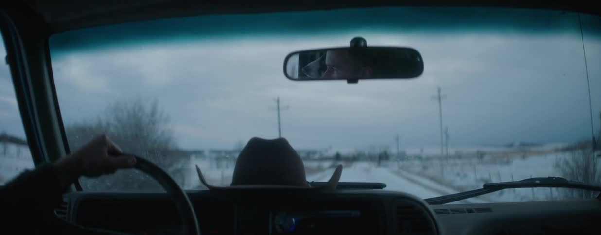 A man's reflection is seen in the rearview mirror as he drives his truck down a snowy country road with his cowboy hat on the dashboard