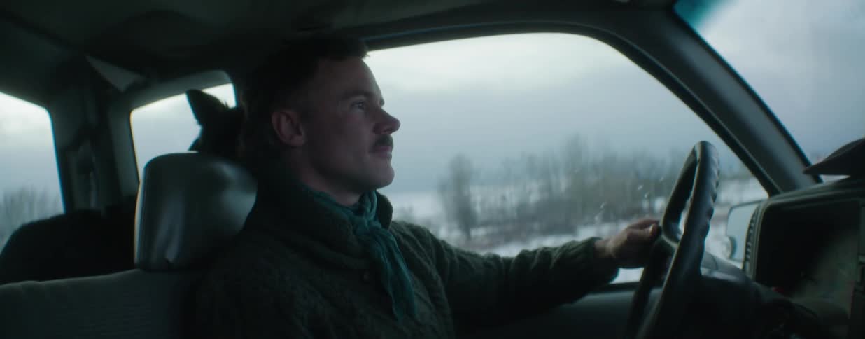 A man with a mustache and his dog in the backseat drives his truck through a serene, snowy rural landscape