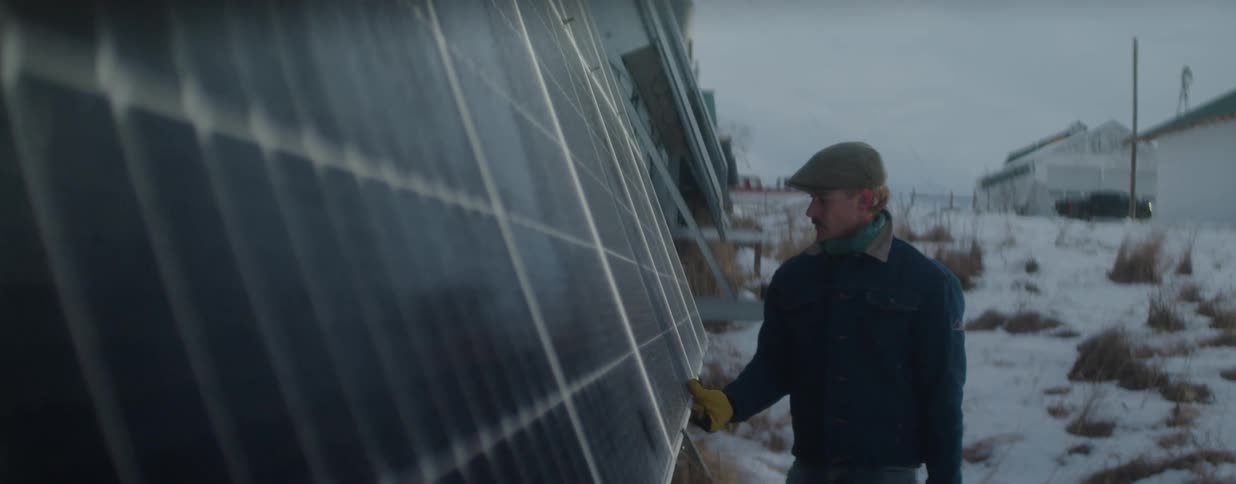 A male technician wearing a cap and gloves inspects a large solar panel array on a rural farm during a cold winter day