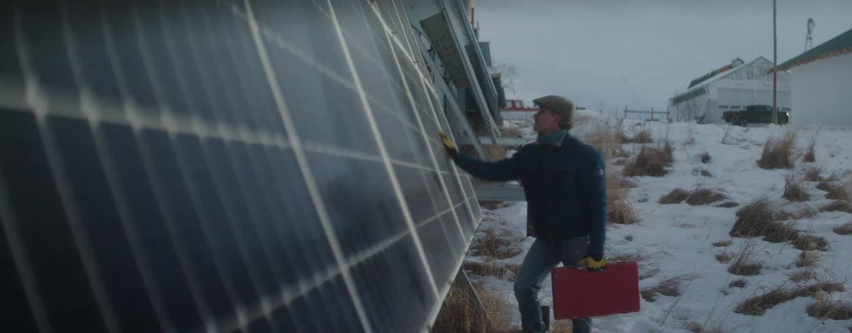 A technician carrying a toolbox inspects a large solar panel array on a snowy, rural property