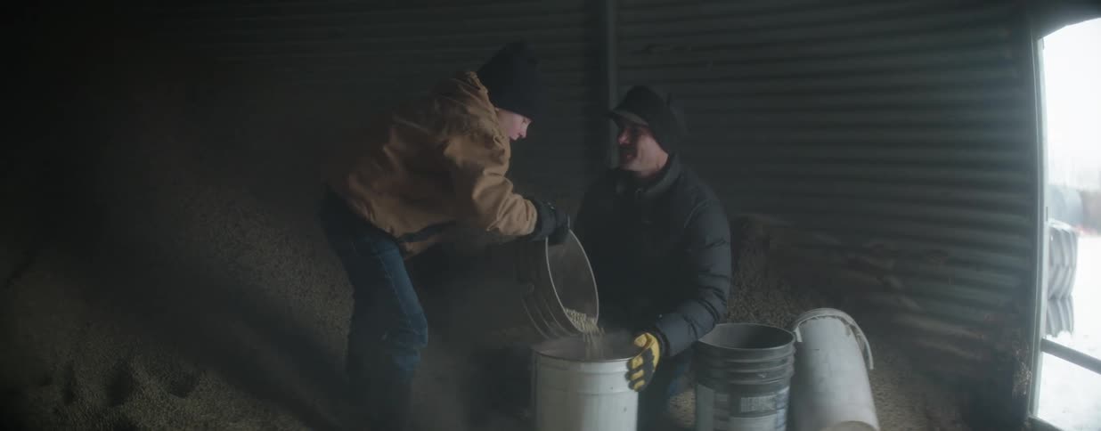 A father and son happily work together in a dusty grain silo, scooping steaming feed into a bucket