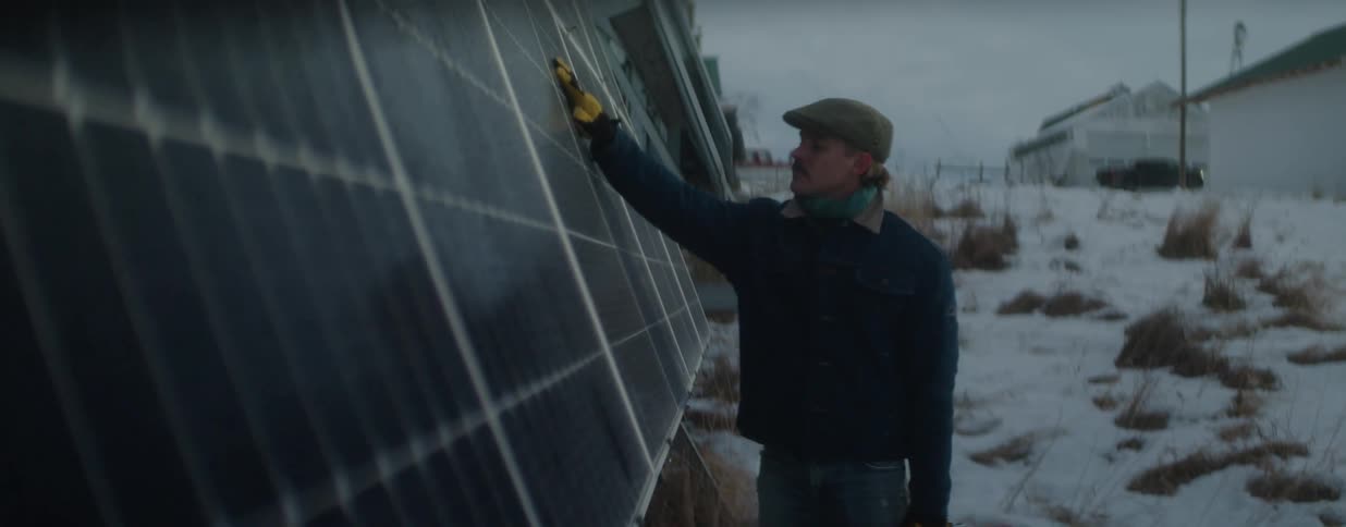 A farmer inspects a row of large solar panels in a snowy field on a cold winter day