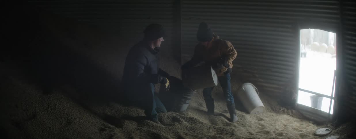 In a dusty, dimly lit grain bin, a father and his young son work together to fill buckets with grain