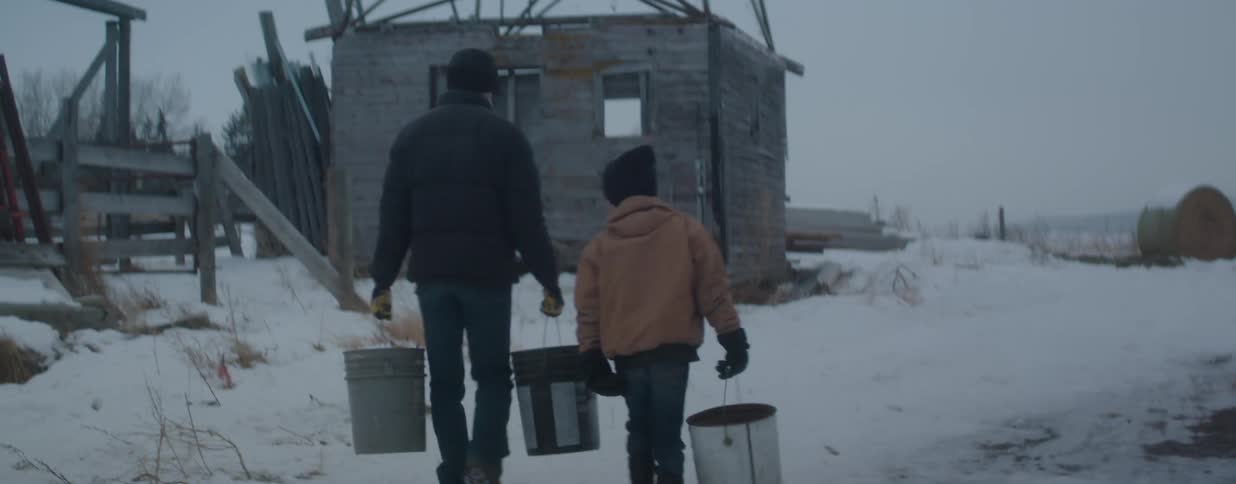 A father and son walk through the snow carrying buckets towards a rustic wooden building on a farm in slow motion