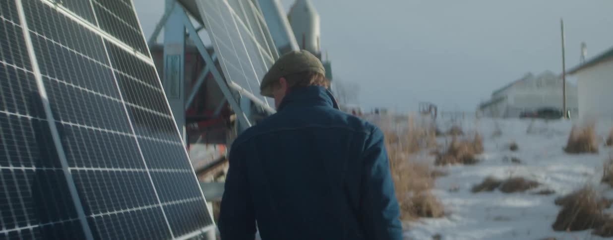 A man wearing a flat cap inspects a large solar panel array in a snowy rural setting during the winter