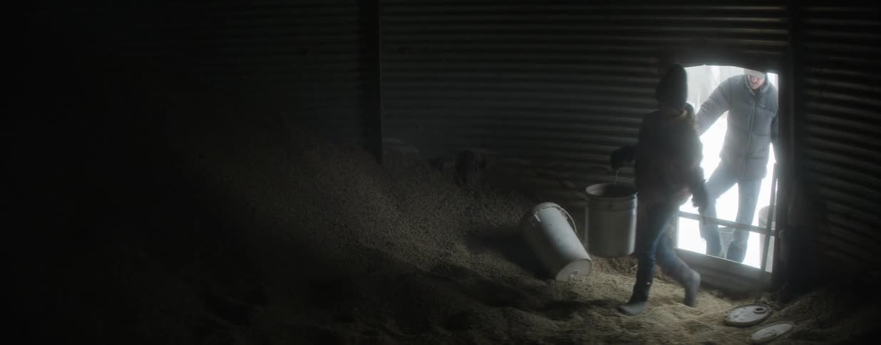 Two farmers scoop feed from a large pile of grain inside a dimly lit silo on a cold, snowy day